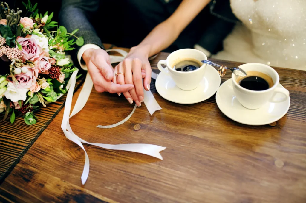 Brautpaar hält sich an den Händen neben zwei Tassen Kaffee und dem Brautstrauß