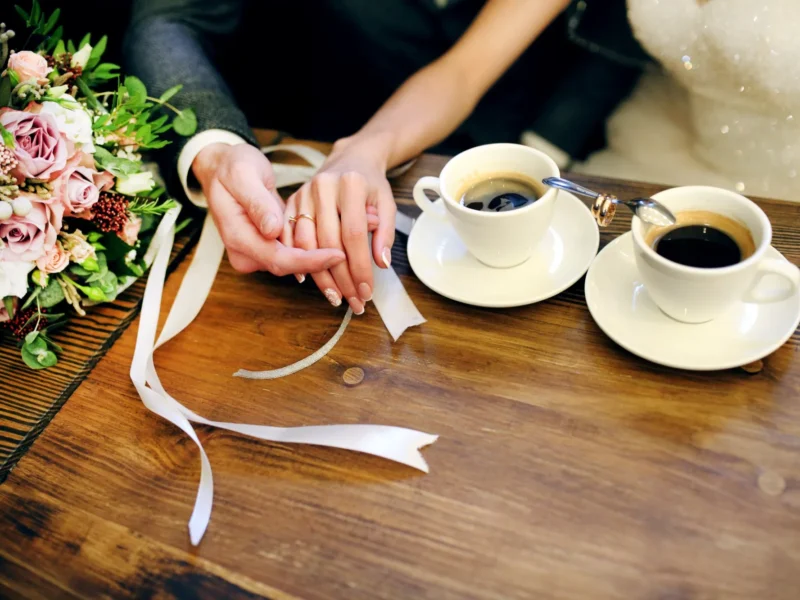 Brautpaar hält sich an den Händen neben zwei Tassen Kaffee und dem Brautstrauß